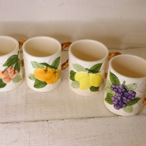 Könnte beinhalten: Set aus vier Keramikbechern mit braunen Griffen. Jeder Becher zeigt ein anderes Fruchtdesign: Kirschen, Orangen, Zitronen und Trauben. Die Becher sind weiß mit grünen Blättern und braunen Ranken.