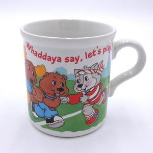May include: White ceramic mug featuring a cartoon illustration of animals playing sports. The mug has the text "Whaddaya say, let's play" printed on it. The handle is white and curved.