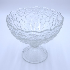 Puede incluir: Un cuenco de pedestal de vidrio transparente con un diseño floral. El cuenco tiene un borde festoneado y una base ancha y redonda.