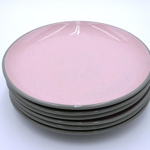 May include: A stack of six pink ceramic plates with a speckled texture. Each plate has a grey rim. The plates are round and appear to be new. The plates are stacked on top of each other.
