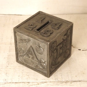 Puede incluir: Una alcancía vintage de metal con forma de cubo con una ranura en la parte superior. Cada lado del cubo presenta una letra y un número diferentes, incluidas las letras A, B y los números 1, 2, 3 y 4.