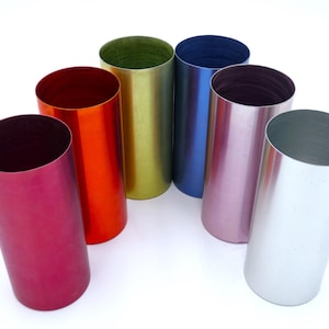 May include: A collection of six cylindrical metal tumblers in a variety of vibrant colors including magenta, orange, lime green, blue, light purple, and silver. Each tumbler features a smooth, solid-colored finish and an open top design.