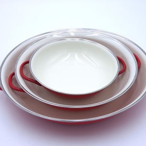 May include: Three nested, red and white enamelware pans with red handles. The pans are oval-shaped and have a silver rim. The interior of the pans is white, and the exterior is red. The pans are arranged in a stack, with the largest pan at the bottom.