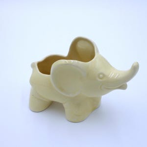 May include: A ceramic elephant planter in a pale yellow color. The planter has a wide opening at the top and a small spout on the side.