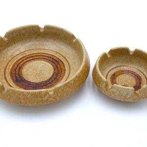 May include: Two beige ceramic ashtrays. The larger ashtray is round with three notches and a brown spiral design in the center. The smaller ashtray has a similar design. Both are on a white background.