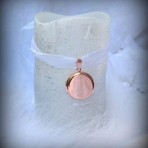 May include: A rose gold locket hangs from a white ribbon wrapped around a textured, off-white candle. The locket is round and has a smooth, reflective surface. The background is soft and blurred, with white feathers.