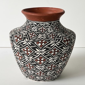 May include: A ceramic vase with a wide, rounded body and a flared rim. The exterior features a repeating geometric pattern of black, white, and terracotta-colored diamonds and cross shapes. The rim is a solid terracotta color.