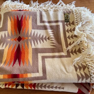 Puede incluir: Manta plegada con un patr&oacute;n geom&eacute;trico en tonos marr&oacute;n, crema, naranja y rojo. La manta tiene un borde con flecos de color crema. El dise&ntilde;o presenta formas de flechas y diamantes.