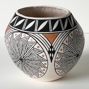 May include: A round ceramic pot featuring a cream-colored base with intricate black geometric patterns and terracotta-colored accents. The design includes circular starburst motifs, a band of leaf-like shapes near the rim, and small dots.