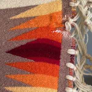 Puede incluir: Primer plano de un textil tejido con un dise&ntilde;o geom&eacute;trico. El patr&oacute;n presenta tri&aacute;ngulos en tonos amarillos, naranjas y rojos sobre un fondo marr&oacute;n y crema. Flecos de macram&eacute; color crema son visibles en el lado derecho.