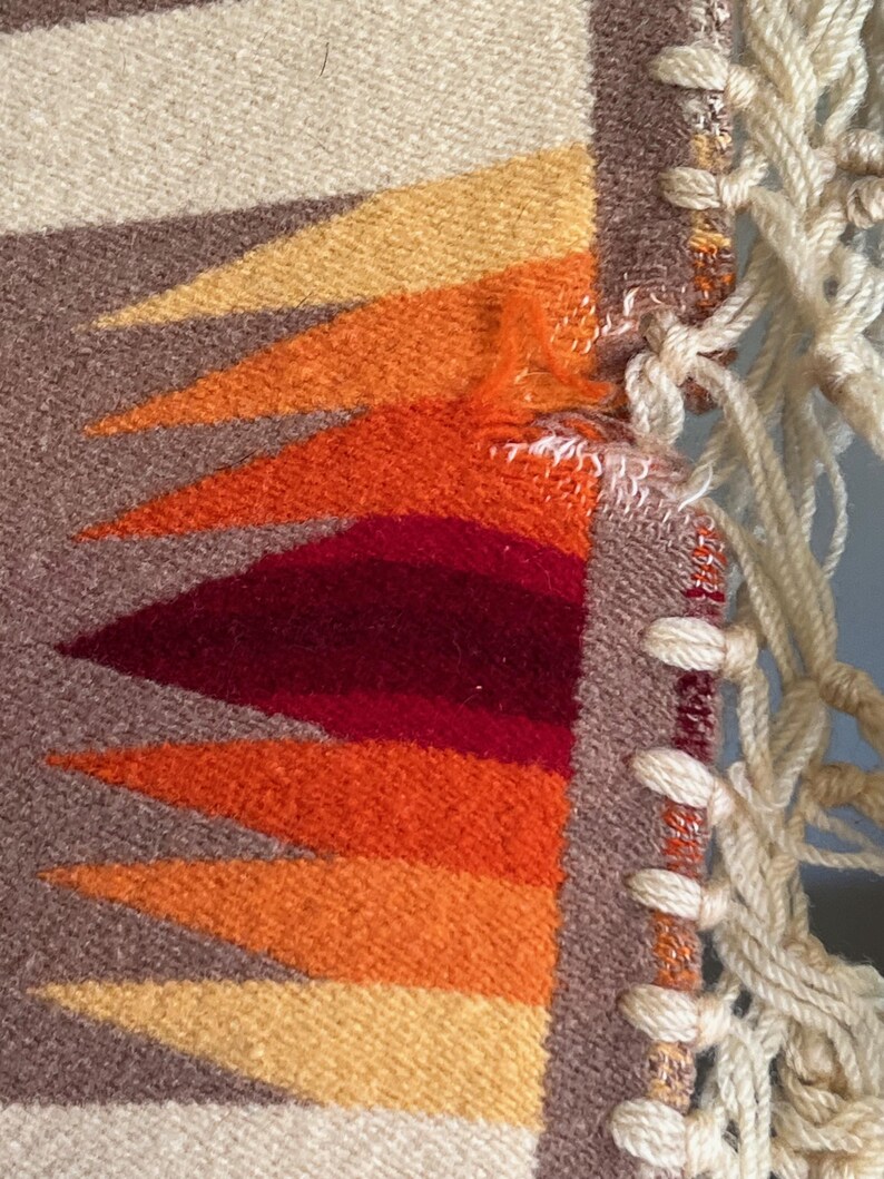 Puede incluir: Primer plano de un textil tejido con un dise&ntilde;o geom&eacute;trico. El patr&oacute;n presenta tri&aacute;ngulos en tonos amarillos, naranjas y rojos sobre un fondo marr&oacute;n y crema. Flecos de macram&eacute; color crema son visibles en el lado derecho.