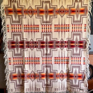 Puede incluir: Manta tejida de color crema con un patr&oacute;n geom&eacute;trico en tonos grises, rojos, naranjas y marrones. La manta presenta un dise&ntilde;o repetitivo en forma de cruz y est&aacute; adornada con flecos en los bordes. Un textil decorativo para la decoraci&oacute;n del hogar.