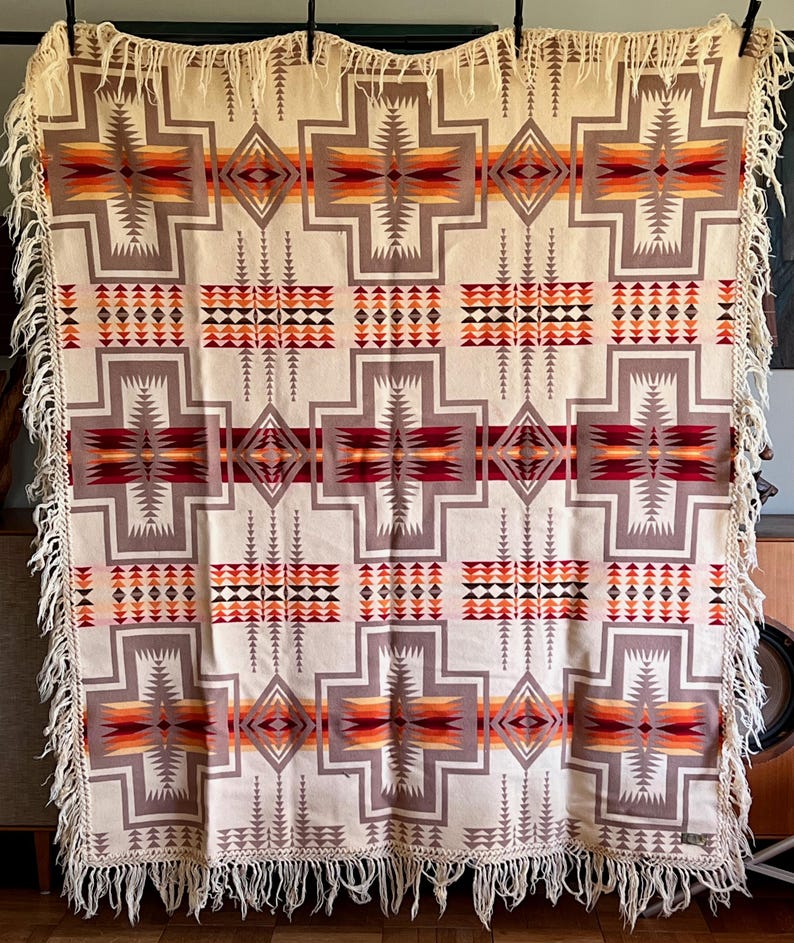 Puede incluir: Manta tejida de color crema con un patr&oacute;n geom&eacute;trico en tonos grises, rojos, naranjas y marrones. La manta presenta un dise&ntilde;o repetitivo en forma de cruz y est&aacute; adornada con flecos en los bordes. Un textil decorativo para la decoraci&oacute;n del hogar.