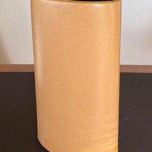 May include: A tall, tan ceramic vase with a slightly oval shape. The vase has a dark blue interior and a textured surface. The vase is approximately 10 inches tall and 4 inches wide.