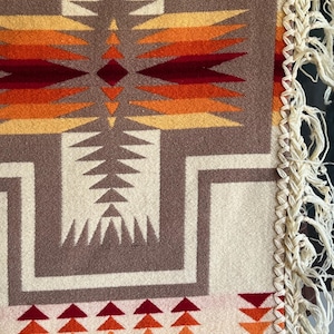 Puede incluir: Primer plano de una manta tejida con un patr&oacute;n geom&eacute;trico en tonos marr&oacute;n, crema, naranja y rojo. El dise&ntilde;o presenta formas repetidas y un borde con flecos. La manta tiene una est&eacute;tica del suroeste o nativa americana.