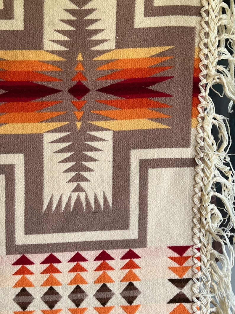 Puede incluir: Primer plano de una manta tejida con un patr&oacute;n geom&eacute;trico en tonos marr&oacute;n, crema, naranja y rojo. El dise&ntilde;o presenta formas repetidas y un borde con flecos. La manta tiene una est&eacute;tica del suroeste o nativa americana.