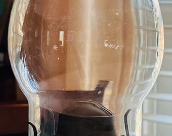 May include: A clear glass oil lamp with a metal burner and a wick. The lamp is sitting on a white surface.