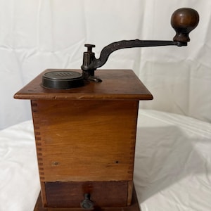 May include: A vintage wooden coffee grinder with a dark metal handle and a small drawer. The grinder has a square wooden body with a dark metal top and a wooden handle with a round knob.