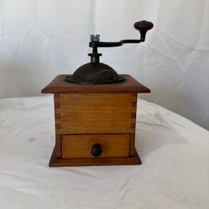 May include: A vintage wooden coffee grinder with a dark metal grinding mechanism and a wooden handle. The grinder has a small drawer and is made of light brown wood. The handle is dark brown. The grinder is on a white surface.