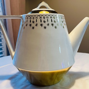 May include: White and gold ceramic teapot with a starburst pattern around the rim. The teapot has a gold base and handle, and a spout. The lid has a gold handle. A classic tea accessory.