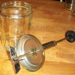 May include: A vintage glass jar with measurement markings sits next to a metal and black hand-operated food mixer. The mixer has a circular base and a blade attachment. The items are on a wooden surface.