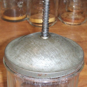 May include: A vintage glass butter churn with a wooden handle and a metal spring mechanism. The churn is made of clear glass and has a silver-colored metal lid.