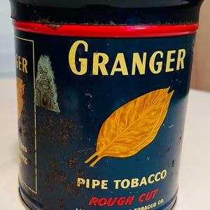 May include: A blue metal tin with a red band around the top. The tin has a yellow label with the text "GRANGER PIPE TOBACCO ROUGH CUT" and the text "LIGGETT & MYERS TOBACCO CO.". The label also features a yellow tobacco leaf.