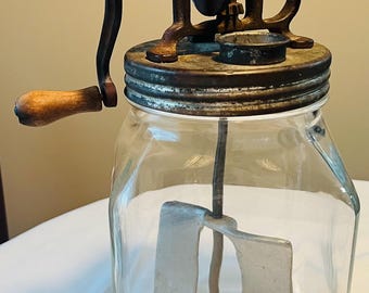 May include: Antique butter churn with a glass jar and a metal crank. The churn has a wooden handle and a metal blade inside the jar.