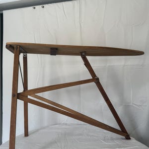 May include: A vintage wooden ironing board with a rectangular top and angled legs. The board is made of light brown wood and features a metal hinge and support. The legs are connected with wooden supports.