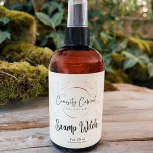 May include: A brown glass bottle with a black spray top and a white label. The label says "Curiosity Cabinet Apothecary" and "Swamp Witch". The bottle contains 8 oz (236 ml) and is for external use only.