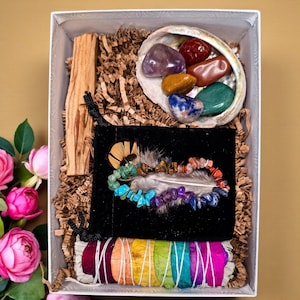 May include: A white box filled with spiritual items. Includes a shell with colorful stones, a black pouch with a feather and beaded bracelet, a bundle of rainbow-colored sage, and a piece of wood. The box is lined with brown paper.
