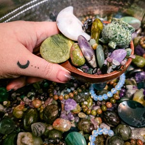 May include: A wooden bowl filled with an assortment of colorful gemstones, including green, purple, and white stones. A carved white moon and a green turtle-shaped stone are visible. The bowl is surrounded by more gemstones and beaded bracelets.