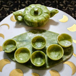 May include: A light green stone tea set consisting of a pumpkin-shaped teapot with a lid and six small matching tea cups. The set is arranged on a matching oval stone tray. The pieces feature a smooth, polished finish with natural veining.