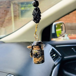 May include: A black and gold car charm with a small glass bottle filled with dried herbs and crystals. The bottle is sealed with black wax and has a gold chain attached to it.