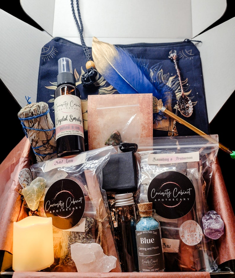 May include: A brown box filled with various items for a spiritual or witchcraft practice. The box contains a white candle, a small bottle of blue liquid, a bag of herbs, a crystal, a feather, a spoon, a sage bundle, and a bottle of crystal smudge. The box is labeled "Curiosity Cabinet Apothecary".