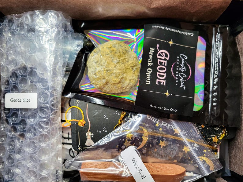 May include: A box filled with various items, including a geode, a wax seal, and a geode slice. The geode is in a clear plastic bag with a label that reads "GEODE Break Open External Use Only". The wax seal is in a clear plastic bag with a label that reads "Wax Seal". The geode slice is in a clear plastic bag with a label that reads "Geode Slice".