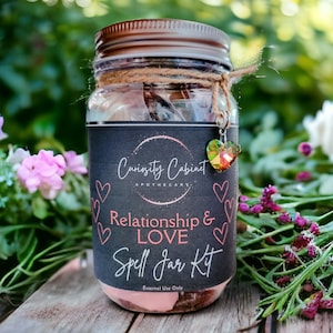 May include: A glass jar with a black label that reads "Curiosity Cabinet Apothecary" and "Relationship & LOVE Spell Jar Kit". The jar is filled with herbs and crystals and has a charm attached to the lid. The jar is sitting on a wooden surface with green foliage in the background.