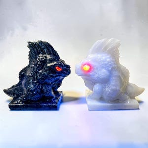 May include: Two small creature figurines, one black and one white, each featuring a prominent dorsal fin, textured scales, and a single glowing eye. The black figurine has a red glowing eye, while the white figurine has a bright yellow glowing eye.