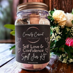 May include: A glass jar with a black label that reads "Curiosity Cabinet Apothecary. Self Love & Confidence Spell Jar Kit. External Use Only." The jar is filled with various herbs, crystals, and other ingredients. The jar is surrounded by pink and white flowers.