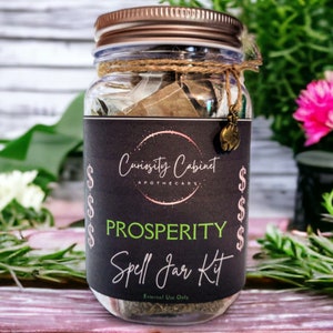 May include: A glass jar with a black label that reads "Curiosity Cabinet Apothecary" and "Prosperity Spell Jar Kit". The jar is filled with herbs and other ingredients. The label also has dollar signs on either side of the text.