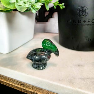 May include: A small, carved stone pelican figurine. The pelican has a green head and beak with a red eye, and a speckled gray body. The figurine is displayed on a white surface.