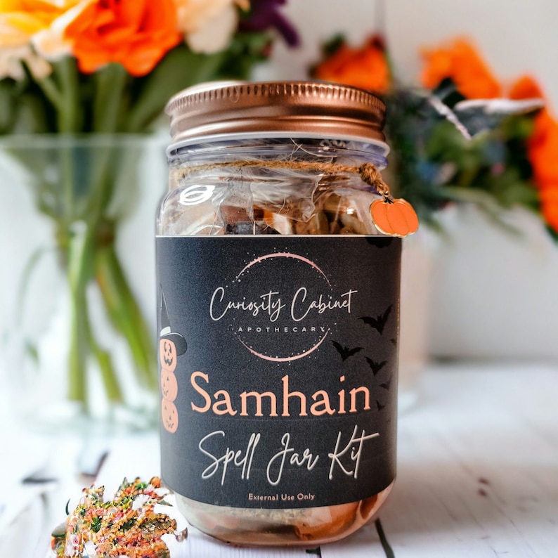 May include: A glass jar with a black label that says "Curiosity Cabinet Apothecary Samhain Spell Jar Kit" and "External Use Only". The jar is filled with various herbs and crystals. The jar has a copper lid and a small orange pumpkin charm.