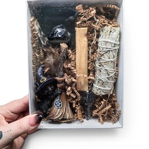 May include: A white box filled with spiritual items, including a black cat figurine with a witch hat, a broom, a crystal, a feather, a palo santo stick, a sage bundle, and a pendulum. The box is filled with brown paper shreds.
