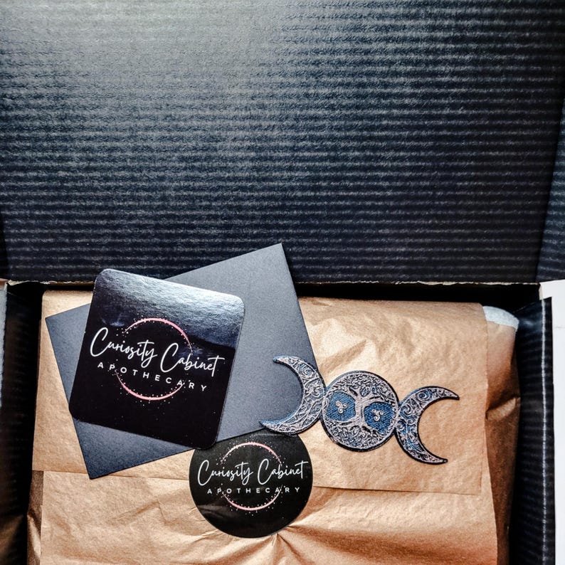 May include: A brown paper package with a black and white sticker that says "Curiosity Cabinet Apothecary". The package also contains a silver triple moon charm with a tree of life design.