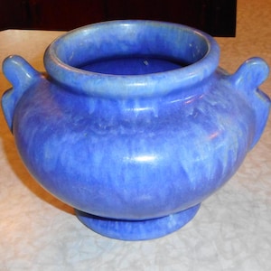 May include: A round ceramic vessel featuring a glossy, mottled blue glaze with a smooth finish. The pot has a wide, open circular mouth, a rounded body, and two small, curved handles on opposite sides, resting on a circular pedestal base.