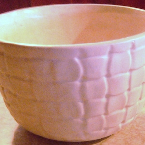 May include: A round, off-white ceramic bowl featuring a raised, embossed basket-weave pattern across its exterior surface. The bowl has a smooth, wide rim and a deep, open interior, showcasing a consistent light cream color throughout the entire piece.
