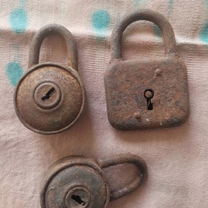 May include: Three rusty metal padlocks with keyholes. The padlocks are different sizes and shapes. The padlocks are on a light brown fabric background.