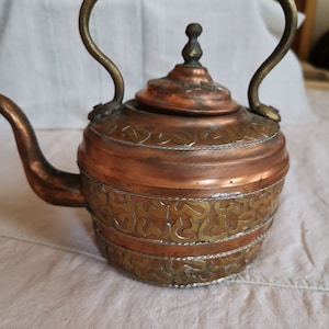 Peut inclure: Théière ancienne en cuivre avec une poignée et des détails en laiton. La théière présente une bande décorative en métal doré autour du corps. Le bec et le couvercle sont également en cuivre, avec un petit bouton en laiton sur le dessus. La poignée est une barre en laiton incurvée.