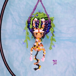 May include: A beaded hanging planter with a small orange and black beaded animal inside. The planter is filled with green and purple beads and has a purple beaded hanger.