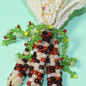 May include: A beaded corn sculpture with three ears of corn in shades of brown, red, and cream. The husks are cream-colored, and green beaded vines are wrapped around the corn. The text "&copy;moontique 2016" is visible.